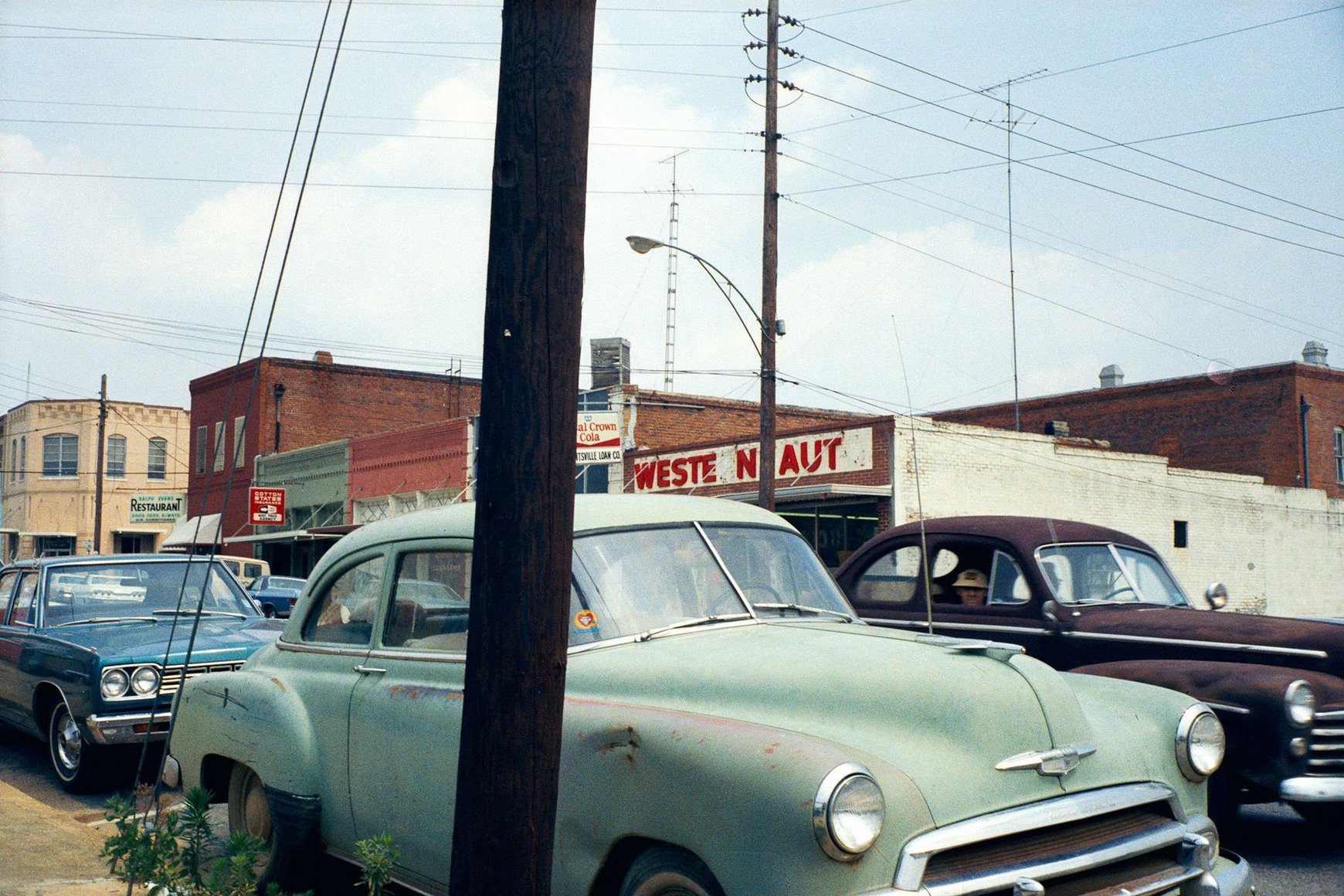 Vintage cars parked on a street in front of storefronts and utility poles.