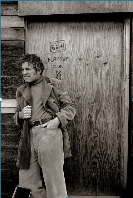 A man with curly hair wearing a coat and turtleneck stands next to a wooden door with graffiti that reads '666' and 'Playboy Club,' with a small Playboy bunny logo. He has one hand in his pocket and the other adjusting his coat.