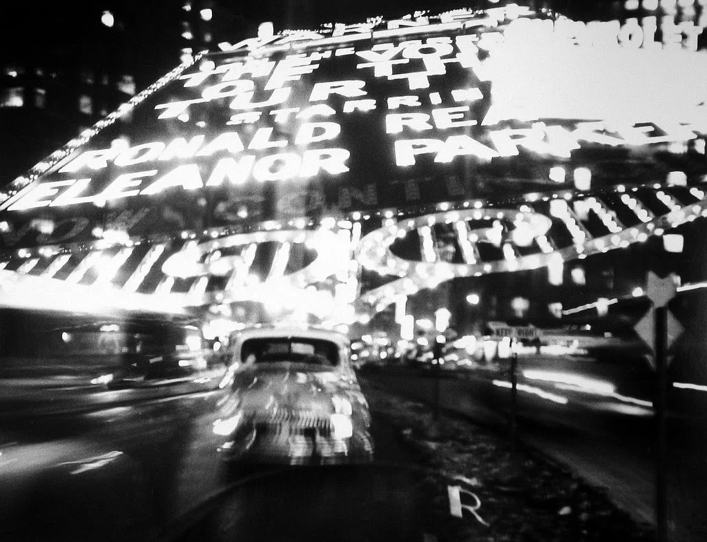 Black and white photo of a street scene at night with a vintage car in the foreground. Brightly lit signs and lights are hanging above the street, creating a lively atmosphere.