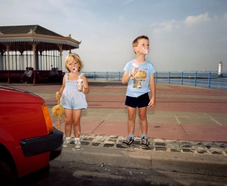 Two young children, a girl and a boy, standing on a seaside promenade with a lighthouse in the background. They are eating ice cream and appear to be enjoying a day out near the ocean.