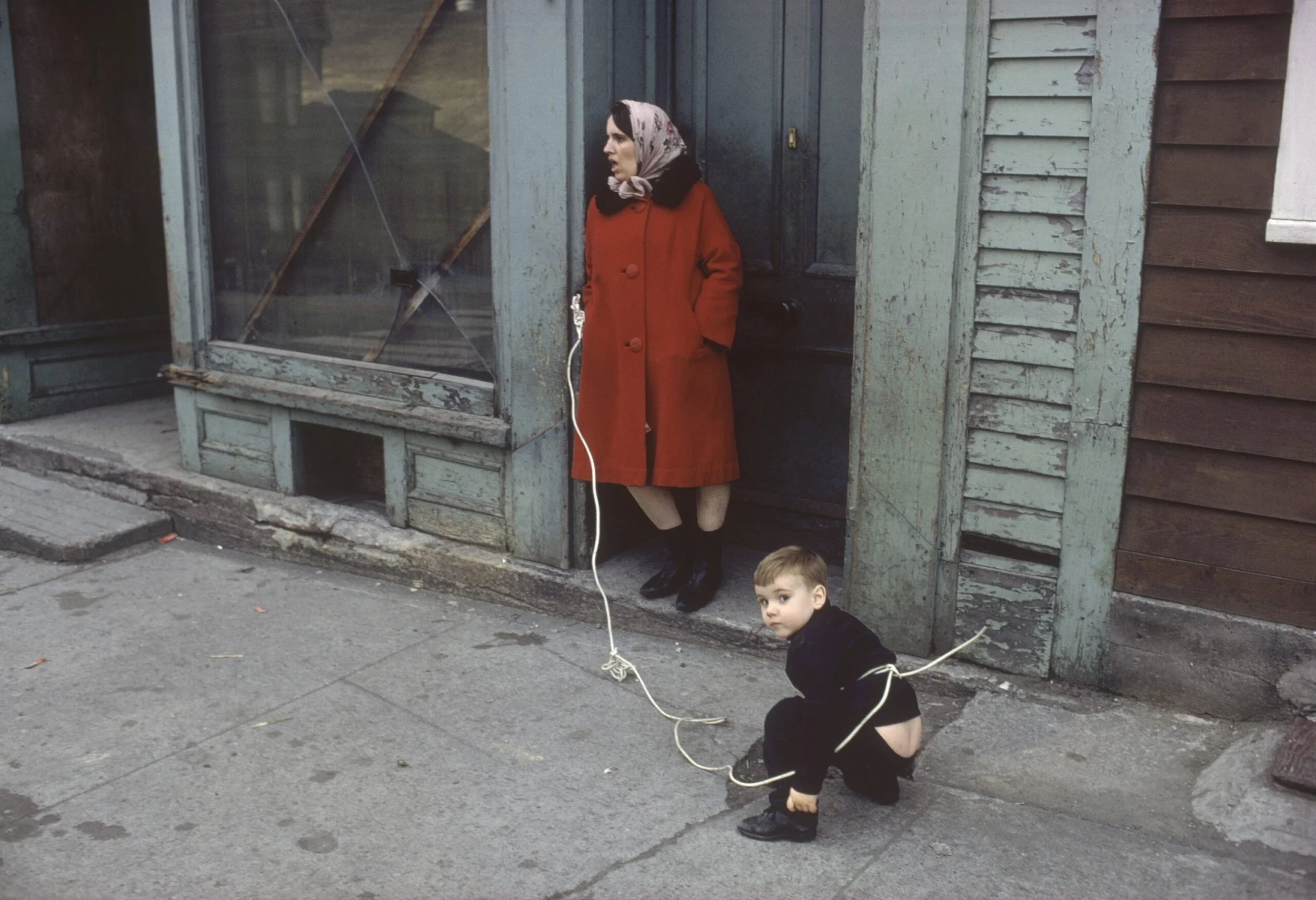 A woman in a red coat and headscarf stands against a weathered building, with a woman in black pants and shoes crouched on the sidewalk in front of her, tied to a longer piece of string, looking at the camera.