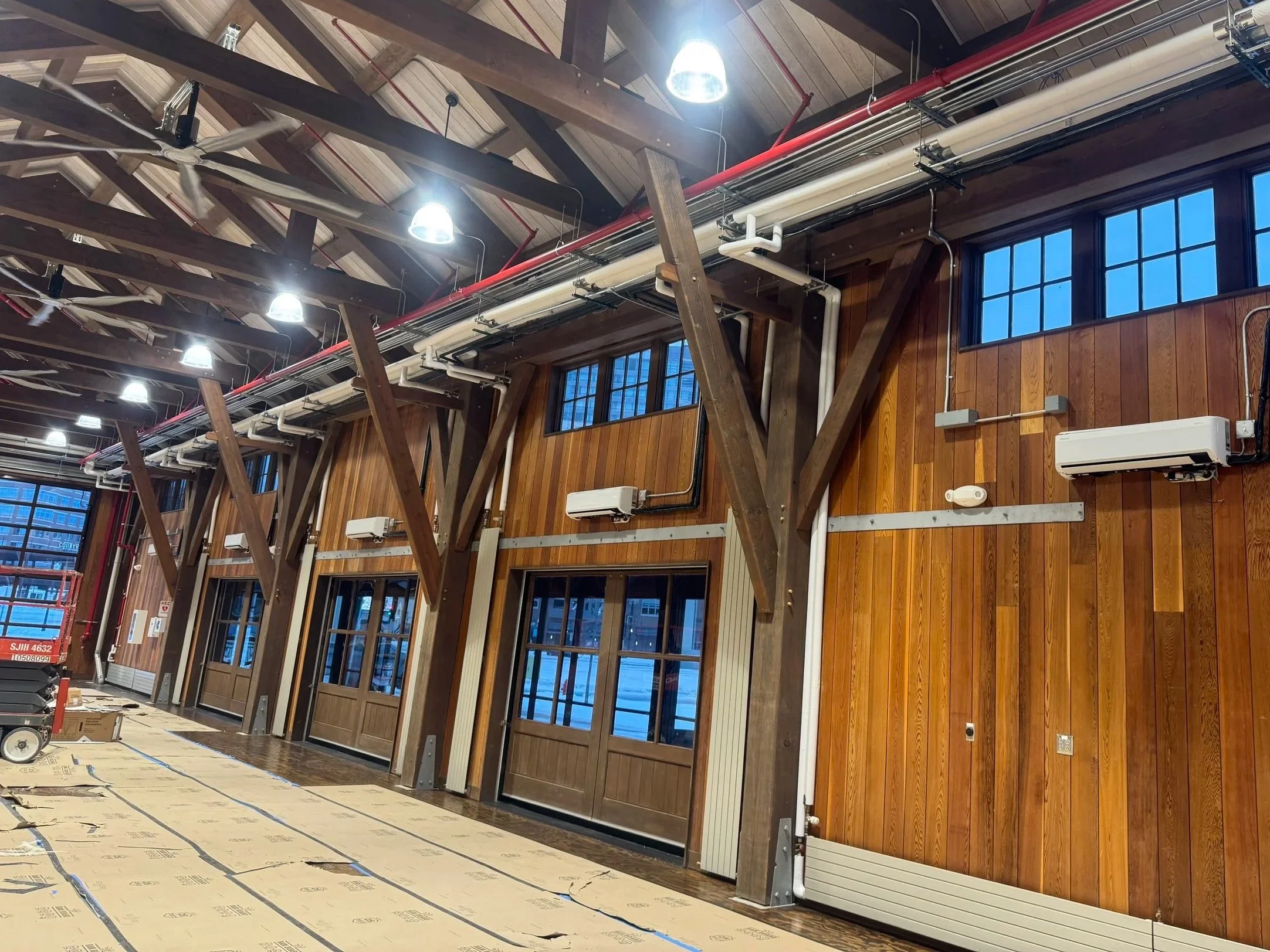 Interior of a building with wooden walls, large windows, exposed ceiling beams, and industrial lighting. Equipment and materials are visible on the floor.
