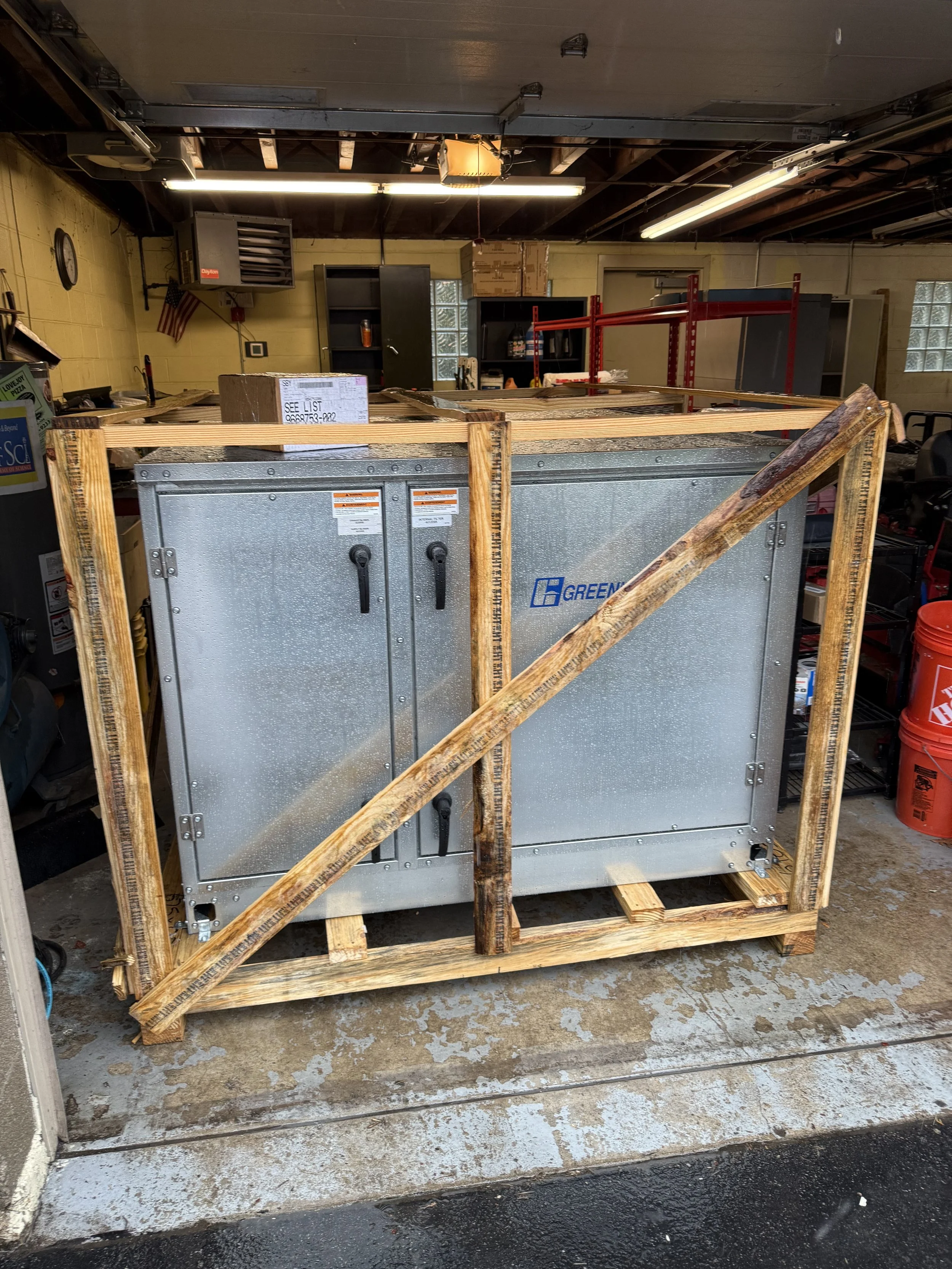 Large industrial HVAC unit labeled Greengair, wrapped in a wooden crate, situated in a workshop with shelves, tools, and equipment in the background.