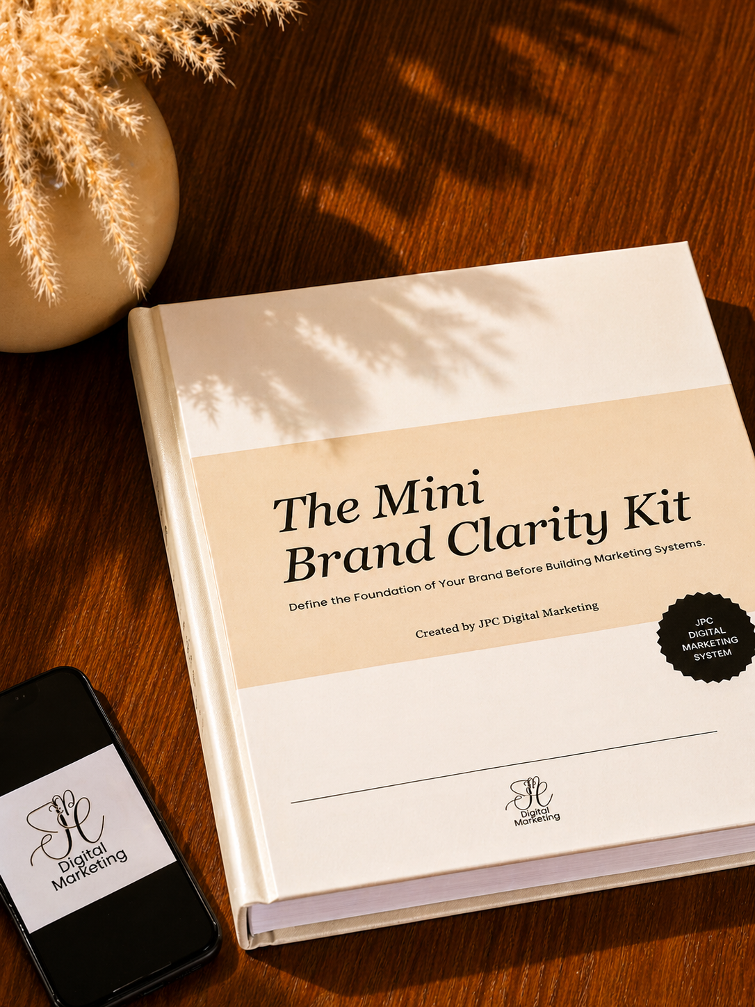 A book titled 'The Mini Brand Clarity Kit' on a wooden table, with a smartphone displaying 'Digital Marketing' logo and a beige vase with dried plants, sunlight casting shadows.