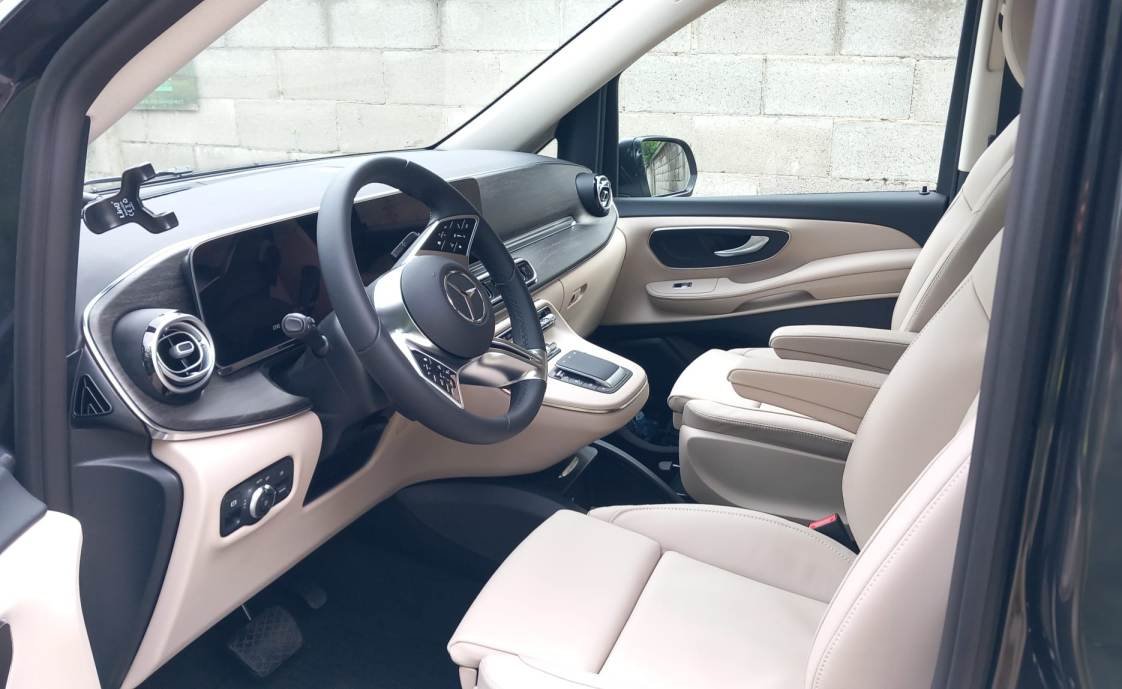 Interno di un'auto con volante, plancia digitale, sedili beige e maniglie sulla porta.