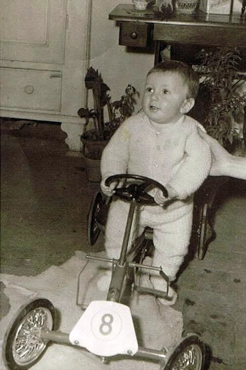 Bambino in abiti comodi che si siede su una go-kart con il numero 8, in un ambiente domestico con mobili e piante sullo sfondo.