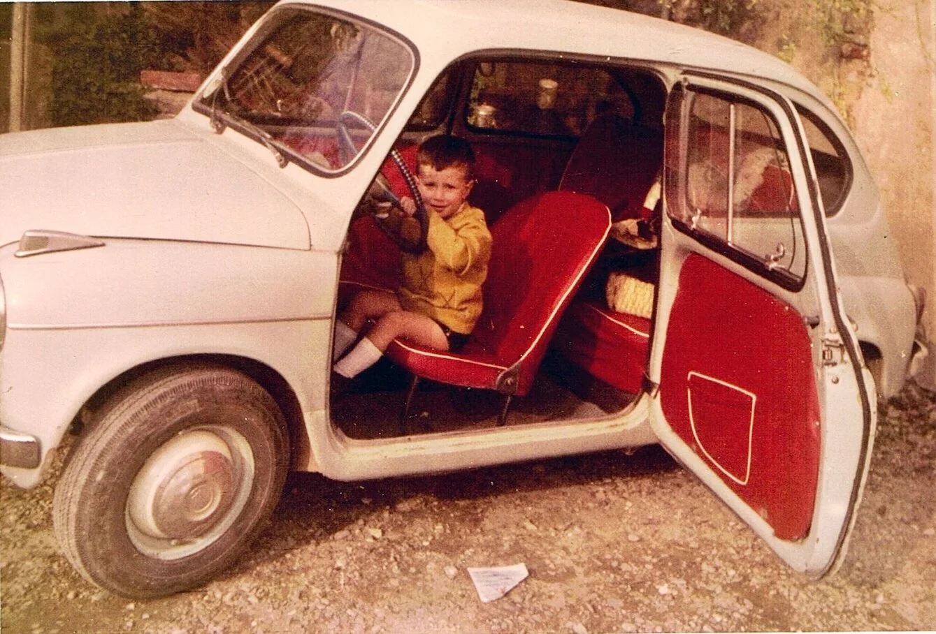 Una bambina seduta sul sedile anteriore di una piccola auto d'epoca con le porte aperte, in un'ambientazione esterna. La vettura è di colore chiaro con interni rosso, e ci sono alcuni oggetti e dettagli all'interno dell'auto.