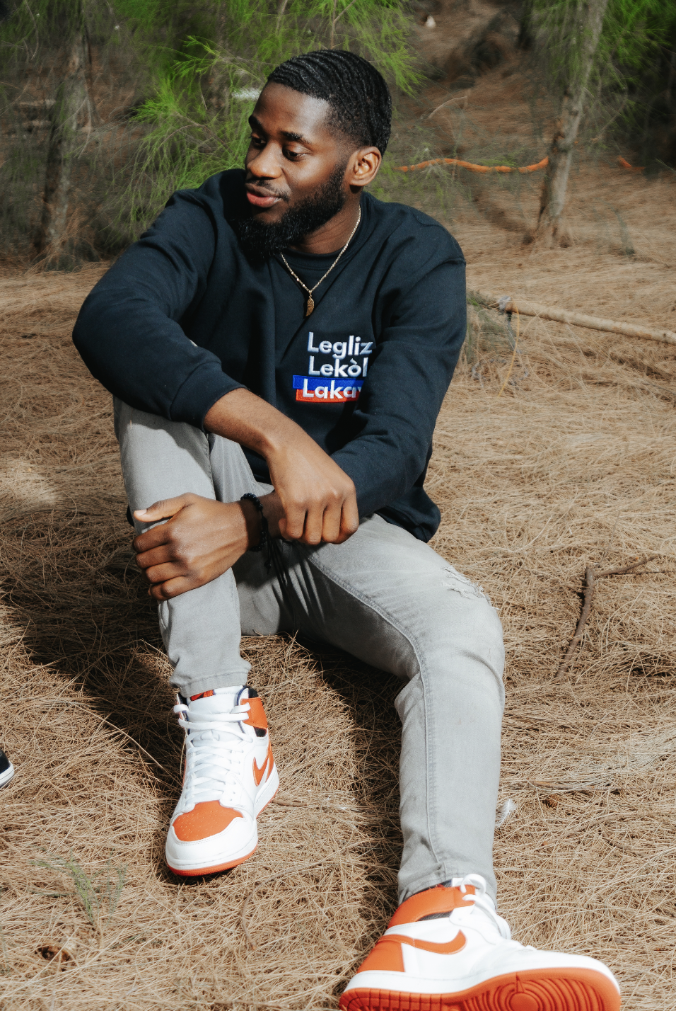 Man sitting outdoors wearing a sweatshirt with text, gray jeans, and red and white sneakers.