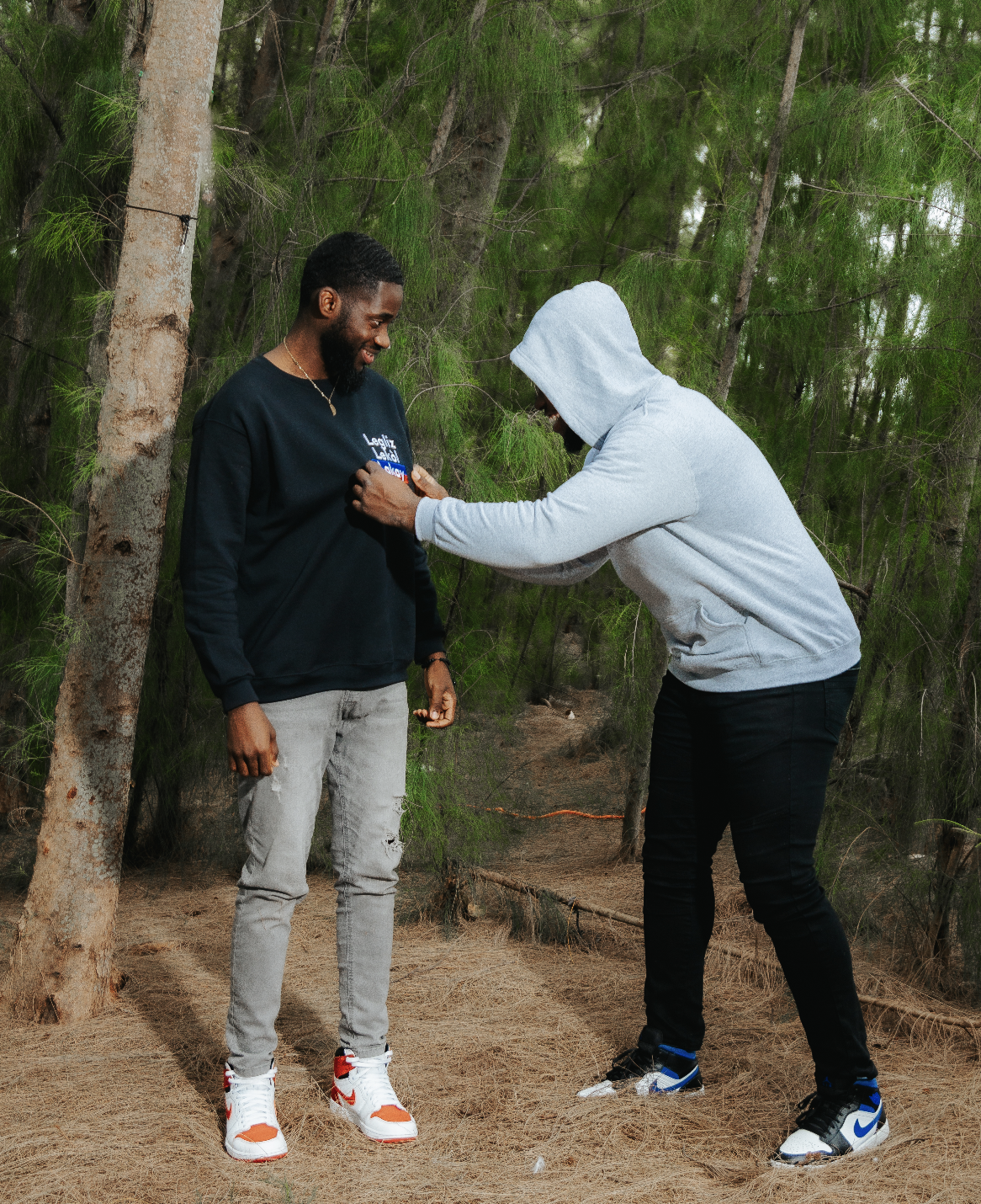 Two people standing outdoors, one wearing a black sweatshirt and ripped jeans, and the other in a gray hoodie and jeans, in a forest setting.