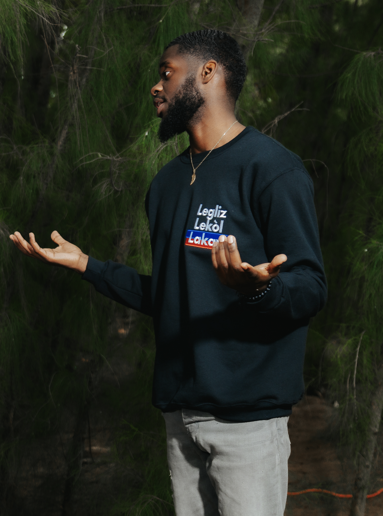 Man wearing a black sweatshirt standing with trees in the background, gesturing with open hands.