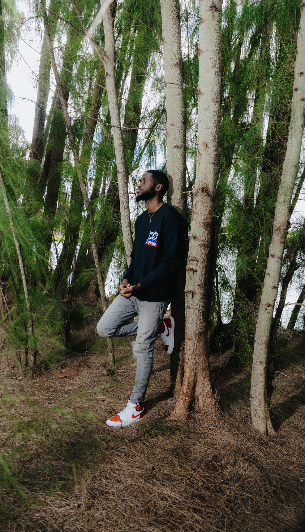 Person in a forest wearing a navy sweater, gray jeans, and white sneakers, leaning against a tree.