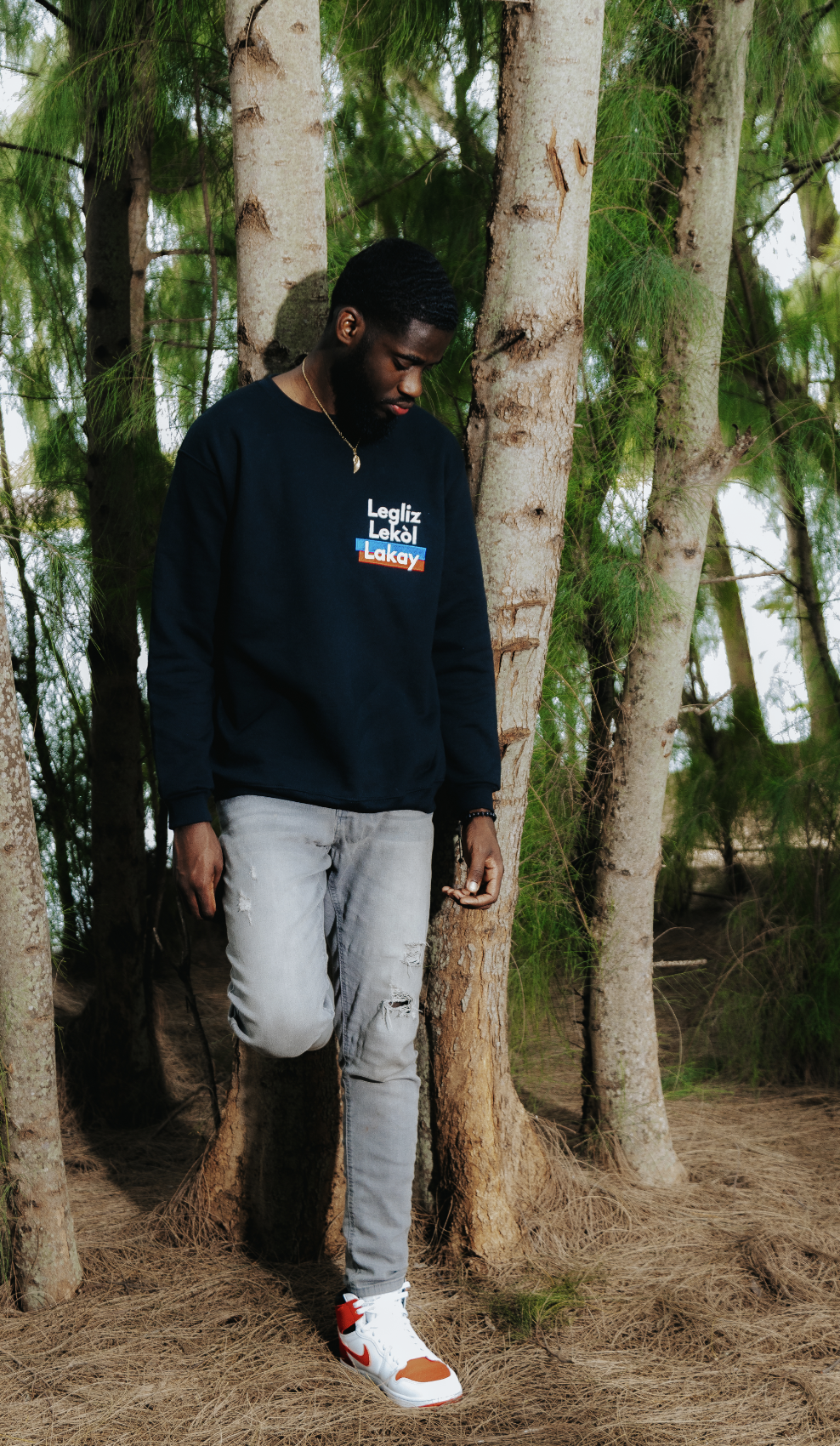 Man in black sweatshirt with text "Legliz Lekòl Lakay," standing in a forest, wearing gray jeans and white sneakers.