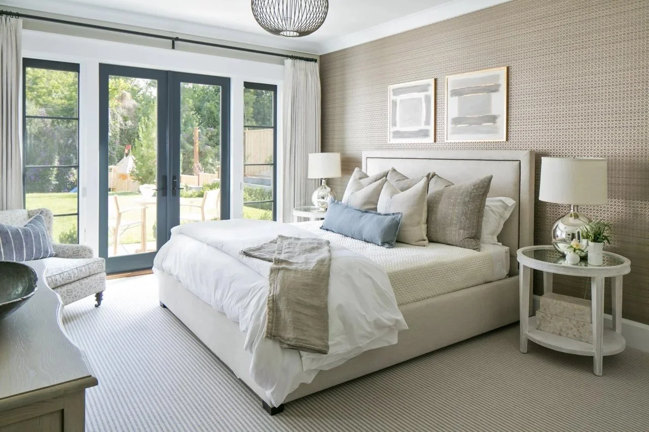 Modern bedroom with neutral decor, featuring a bed with multiple pillows, white bedding, nightstand with a lamp, round table, and large glass doors opening to a garden.