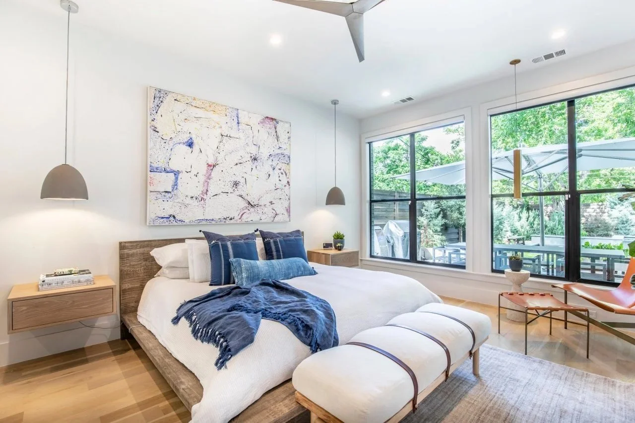 Modern bedroom with wooden bed, blue and white bedding, abstract wall art, large windows, pendant lights, and outdoor view.