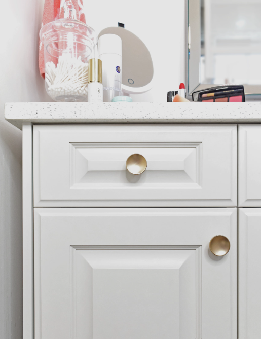 White bathroom vanity with gold knobs, makeup products, a mirror, a jar with cotton swabs, and a pink towel on the counter.