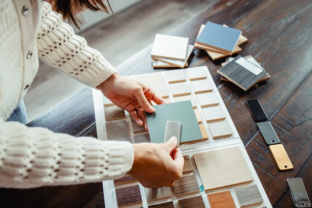 Hands holding up a metal sample to a wood sample block with many other color samples behind.