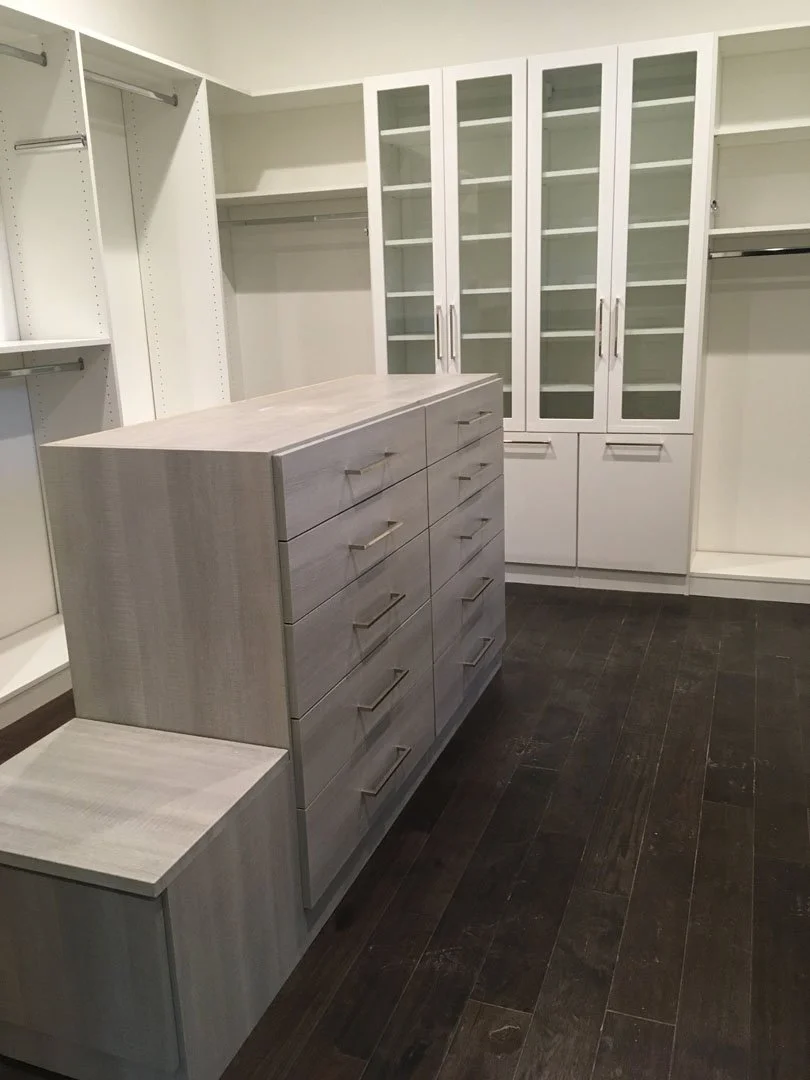 Modern walk-in closet with wooden drawers, glass cabinets, and hanging rods on dark hardwood flooring.
