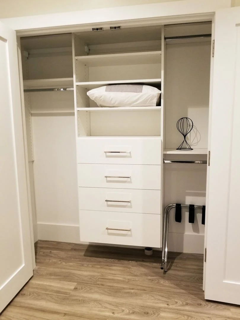 Interior of a white closet with open doors, containing shelves with a pillow, drawers, and a metal sculpture on a shelf.