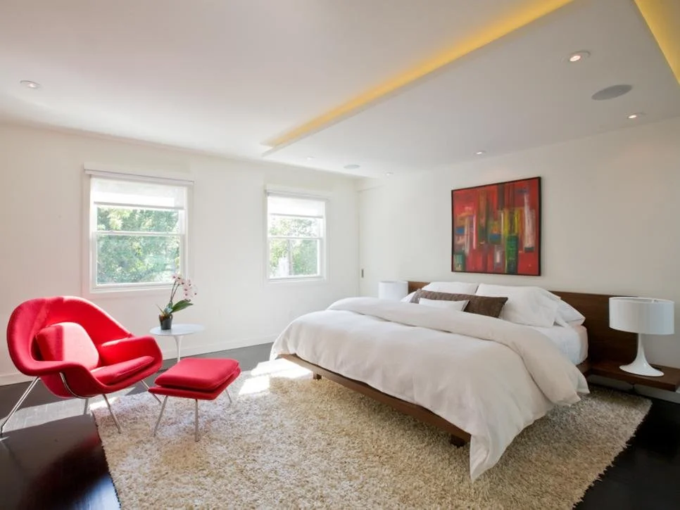 Modern bedroom with a white bed, abstract wall art, red chair and ottoman, white lamps, two windows, and fluffy rug.