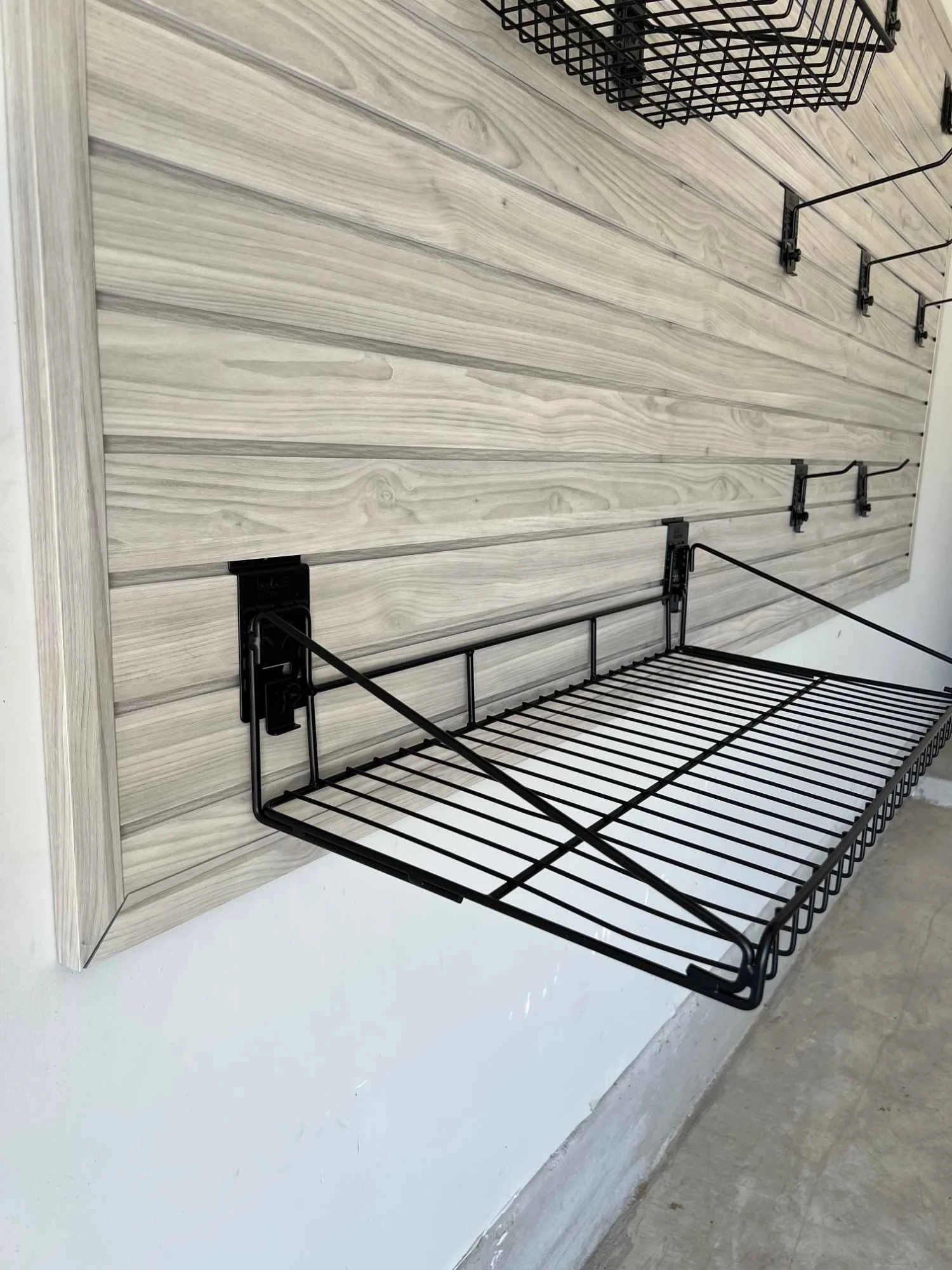 Light gray wood-grain garage slat wall with a black wire shelf, basket, and hooks