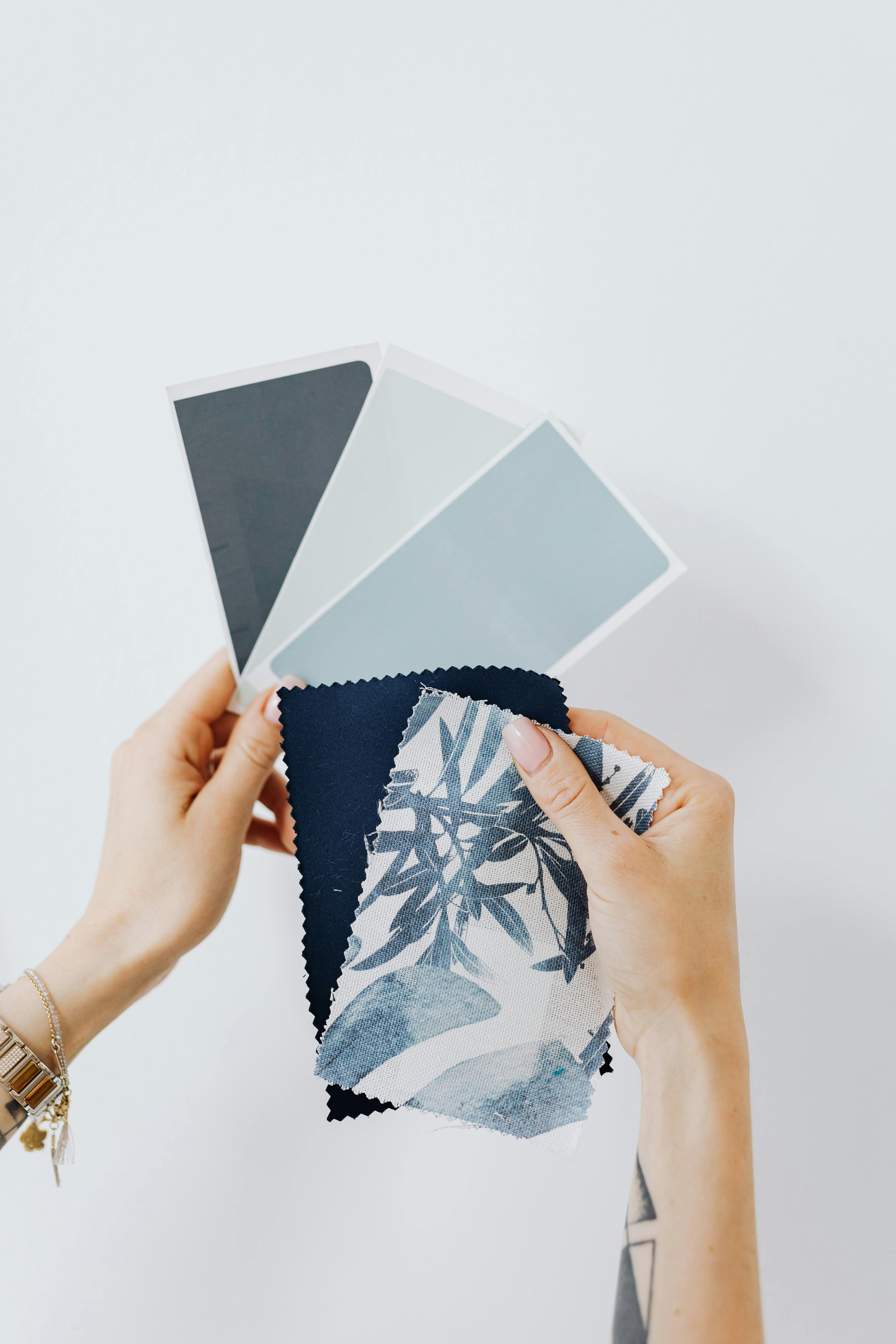 Womans hands holding swatches. The left hand holds 3 matching hues of paint swatches, dark, light and medium blue while the right hand overlays 2 matching fabric or wall paper swatches.