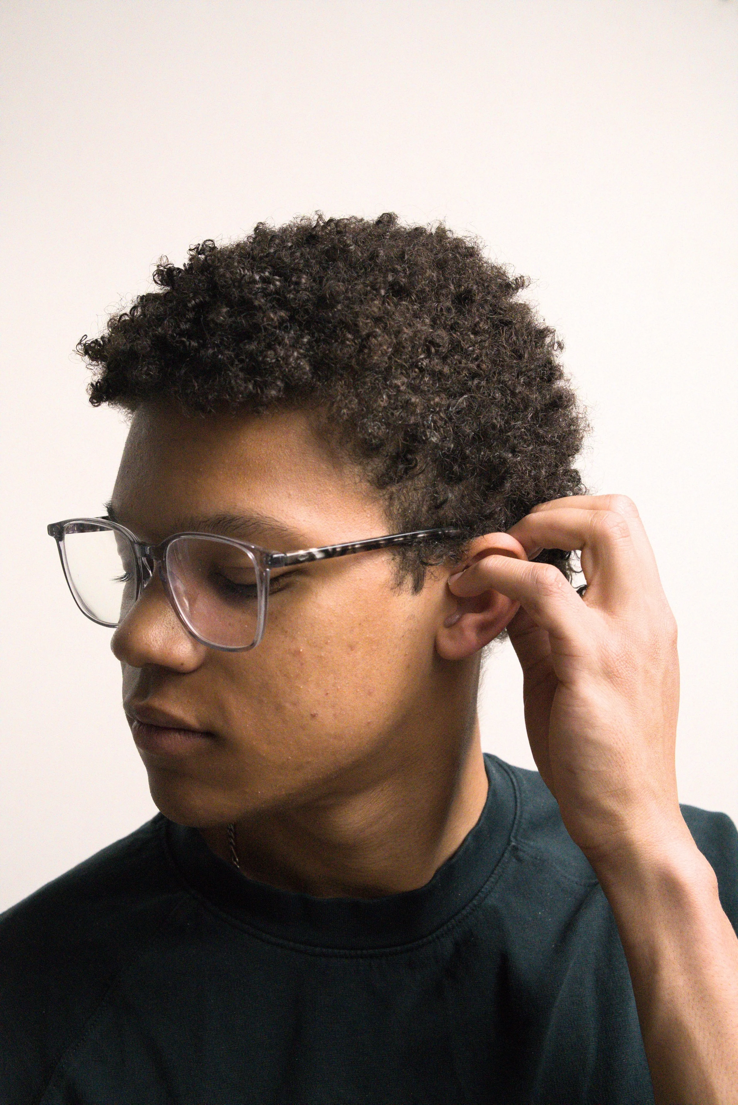 Close-up of a woman with short curly hair, wearing glasses, touching her ear, and wearing a black shirt against a plain white background.