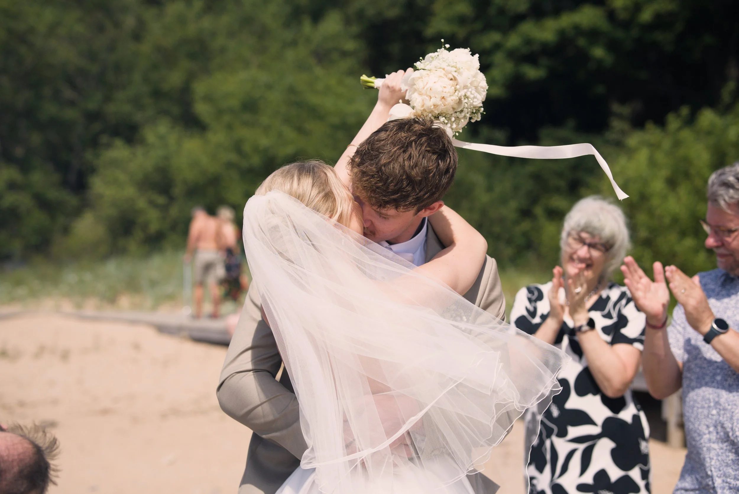 A couple is embracing and kissing at their outdoor wedding, with the bride raising a bouquet of white flowers in the air. Guests are clapping and smiling in the background, set against a green, wooded outdoor setting.