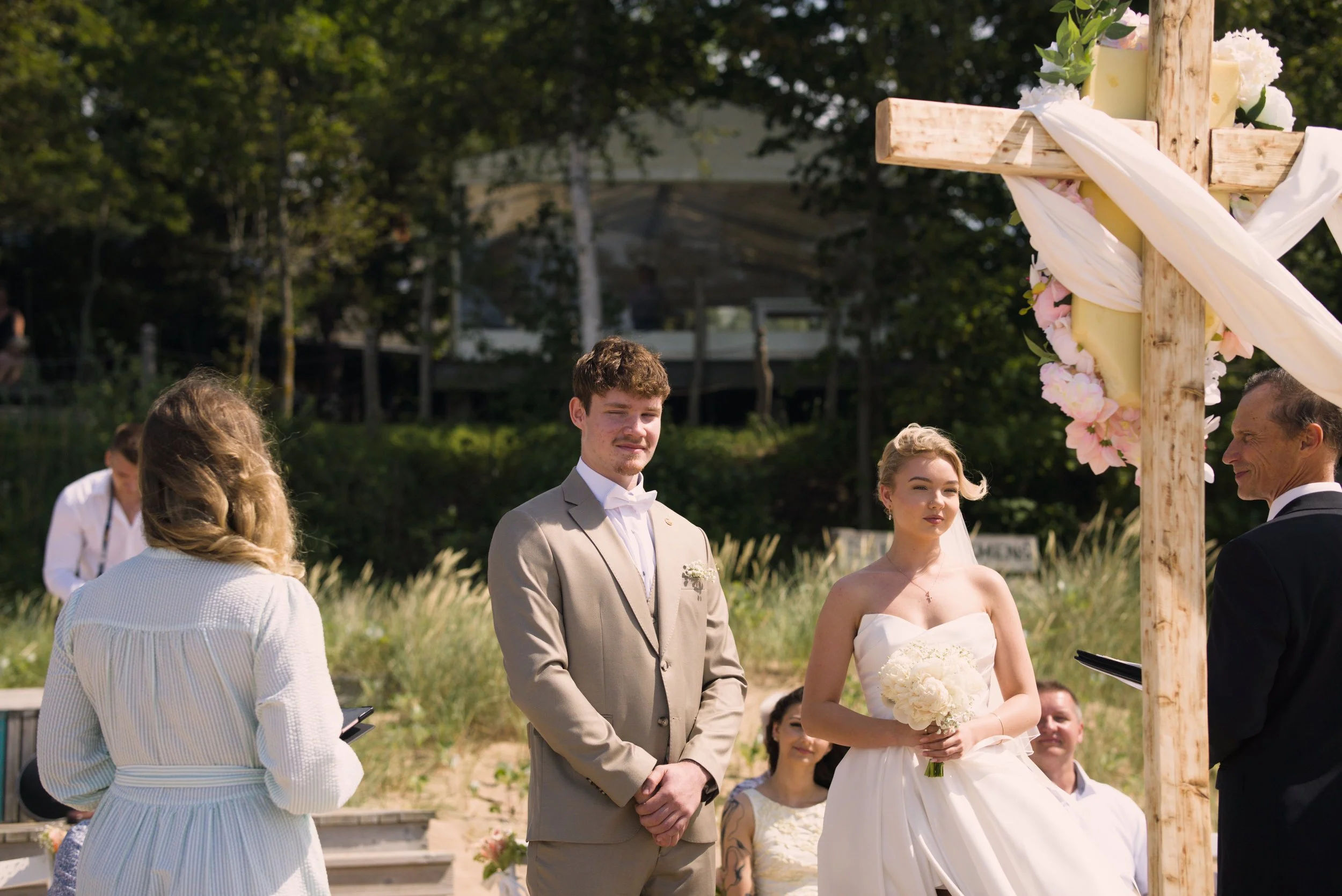 A wedding ceremony outdoors with a bride and groom standing under a wooden cross decorated with white and pink flowers, surrounded by guests.