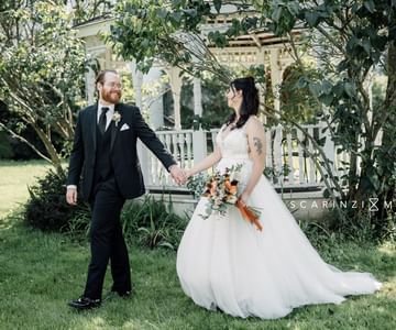 A bride and groom holding hands and smiling outdoors on a grassy area, with trees and a white house in the background.