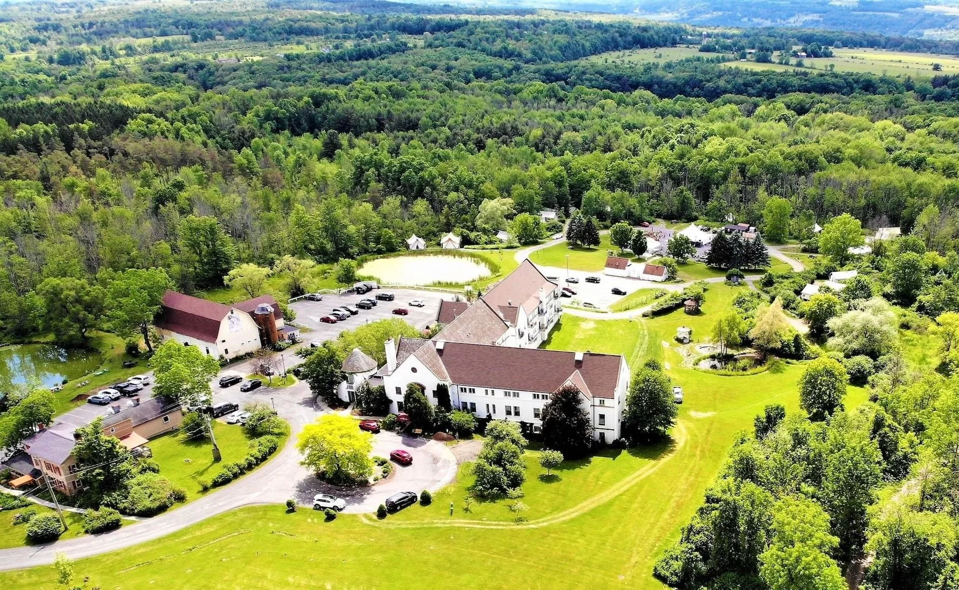 An aerial view of a rural area with a large white church, several houses, parking lots filled with cars, and lush green trees and fields surrounding the buildings.
