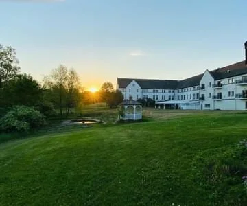 Sunset over a grassy lawn with white residential buildings and a small greenhouse or garden structure in the background.