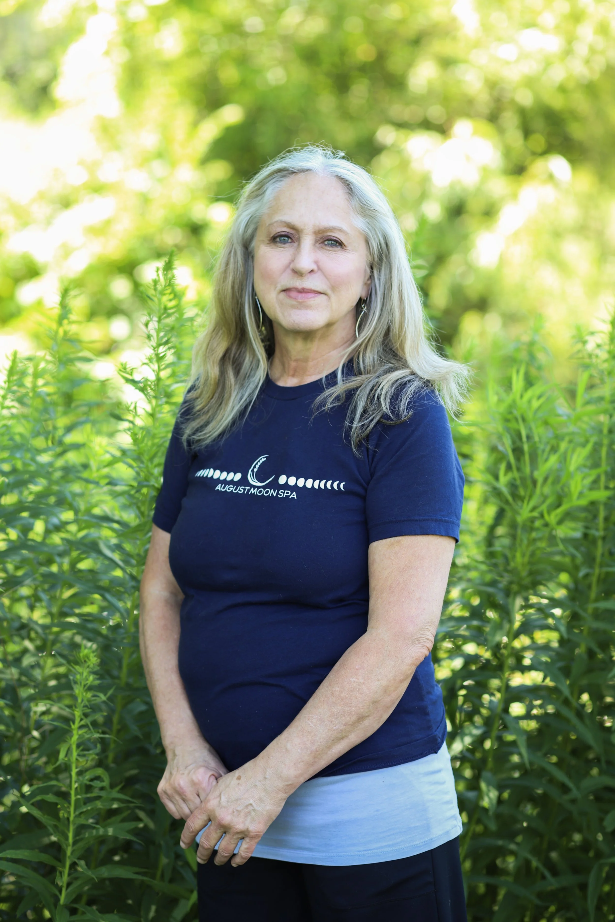 A woman with long blonde hair smiling outdoors with a blurred background of trees.