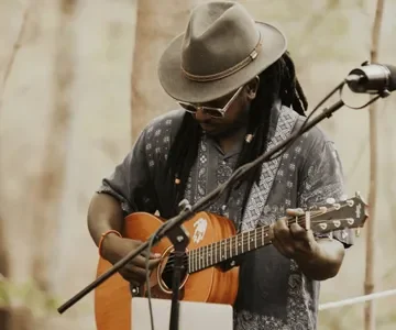 Musician playing an acoustic guitar outdoors, wearing a hat and sunglasses.