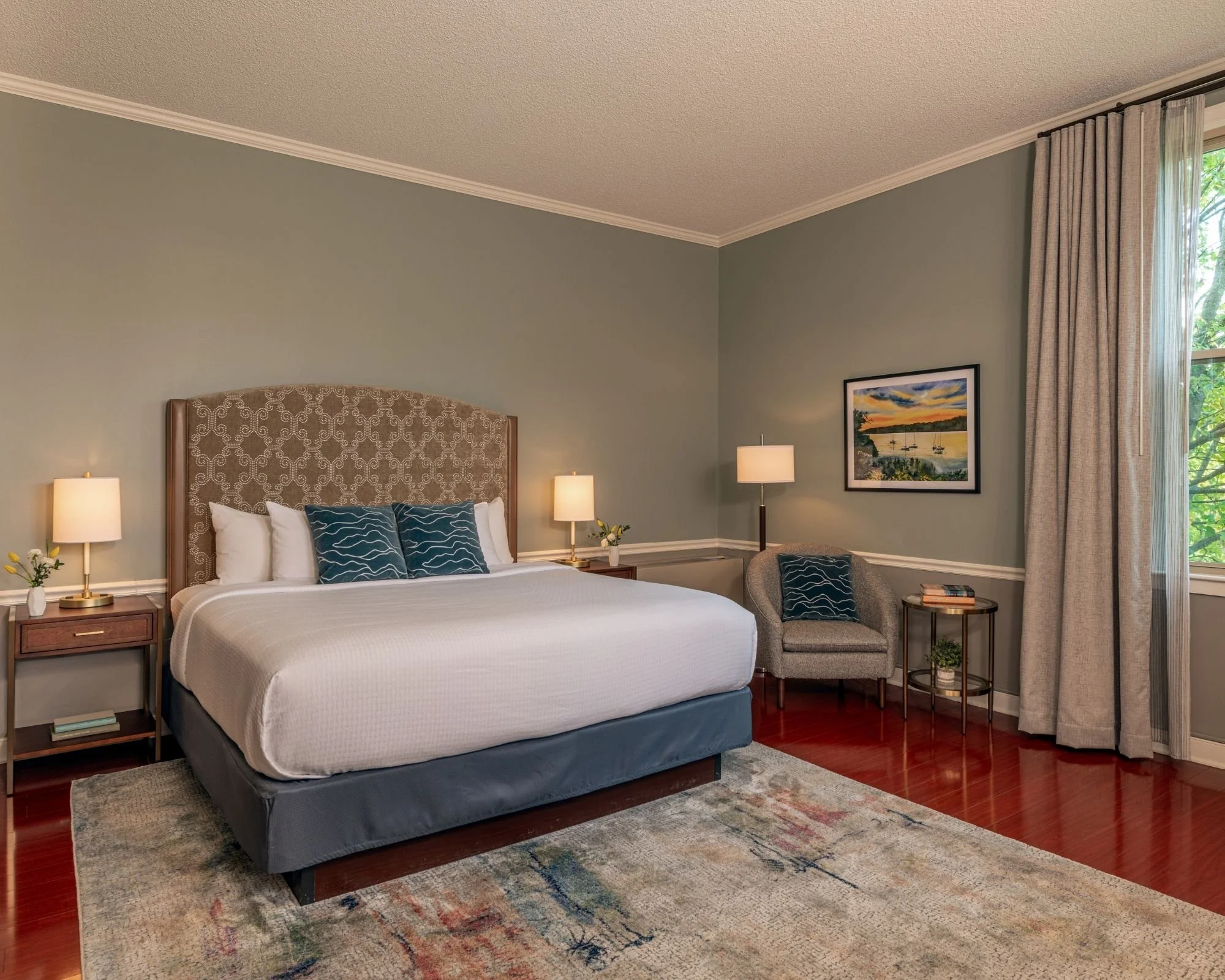 A bedroom with a bed featuring a patterned beige headboard, white bedding, blue cushions with wave patterns, and matching bedside lamps. There is a small nightstand, a picture of a sunset with boats on the wall, a window with curtains, a chair with a cushion, a side table with books, and a patterned area rug on wooden floor.