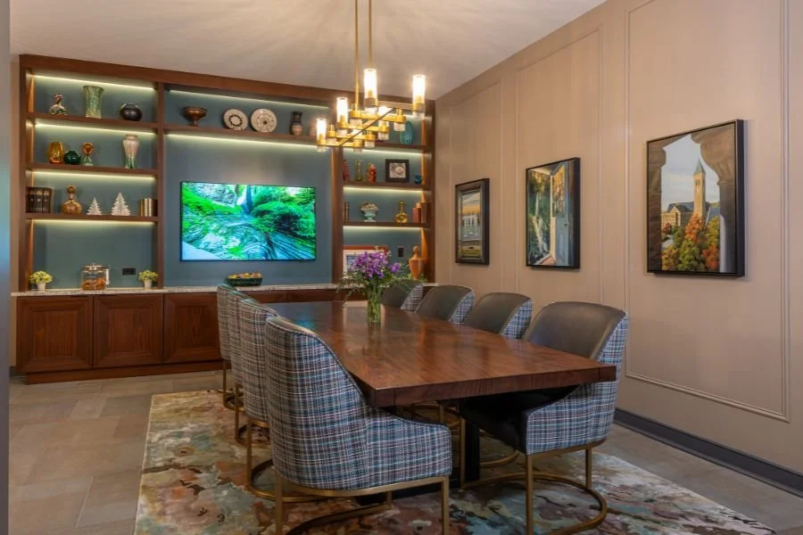 A dining room with a wooden table and eight plaid upholstered chairs, a built-in shelf with decorative items, a wall-mounted TV, framed artwork, a chandelier, and a floral centerpiece.