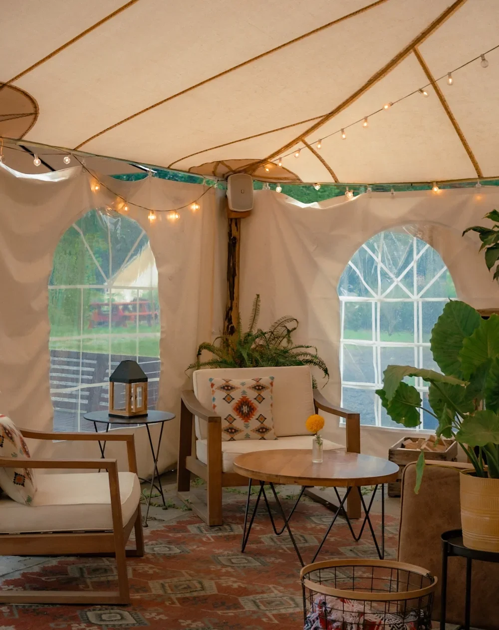 A cozy indoor setup inside a tent with string lights hanging across the ceiling. There are two windows with arched tops, a small round coffee table with a yellow flower in a vase, a white armchair with a colorful pillow, a wooden-framed sofa with a matching pillow, and large green plants. The decor includes a lantern on a side table and a patterned rug on the floor.
