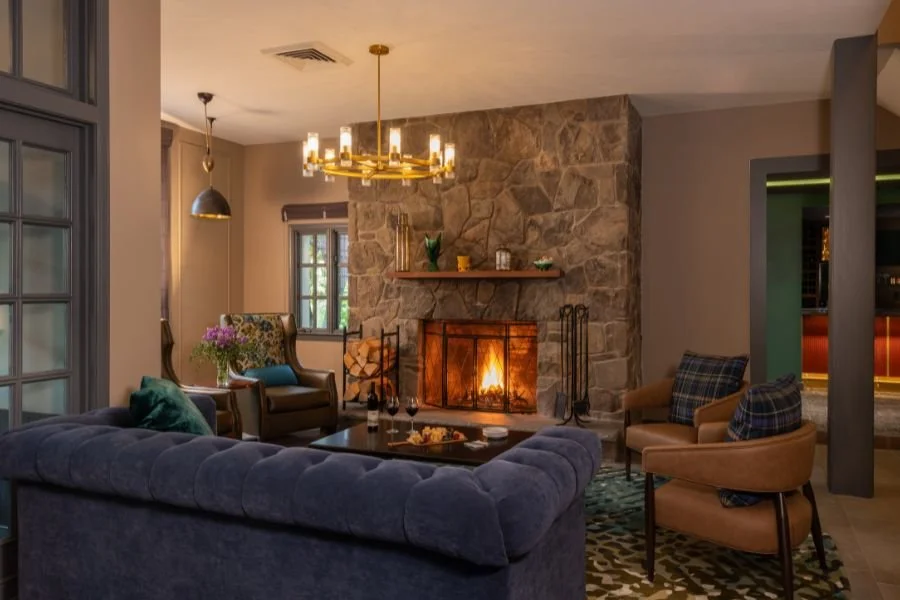 Living room with a gray velvet sofa, a stone fireplace with burning fire, a wooden mantel with decorative items, tan armchairs with plaid pillows, a modern chandelier, and a large window.