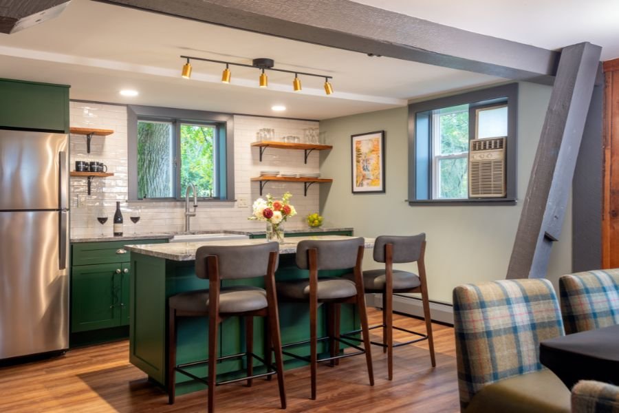 Kitchen with green cabinets, a stainless steel refrigerator, a white tile backsplash, and a window with greenery outside. There is a kitchen island with a vase of flowers and four chairs, and two open wooden shelves on the wall. An air conditioner is installed in the second window.