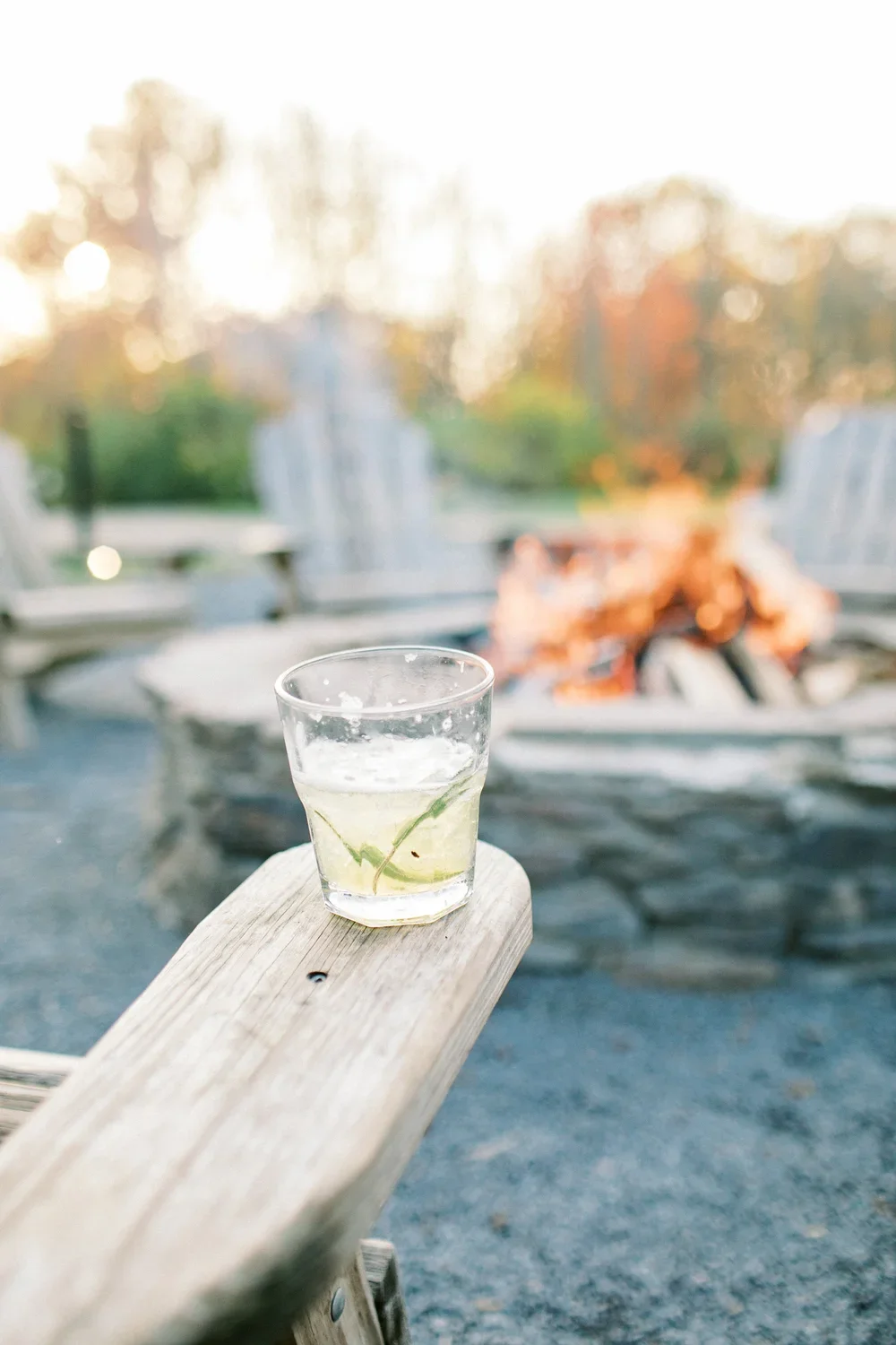 A glass with a lime wedge on a wooden bench outdoors, with a blurred background of chairs and a fire pit.