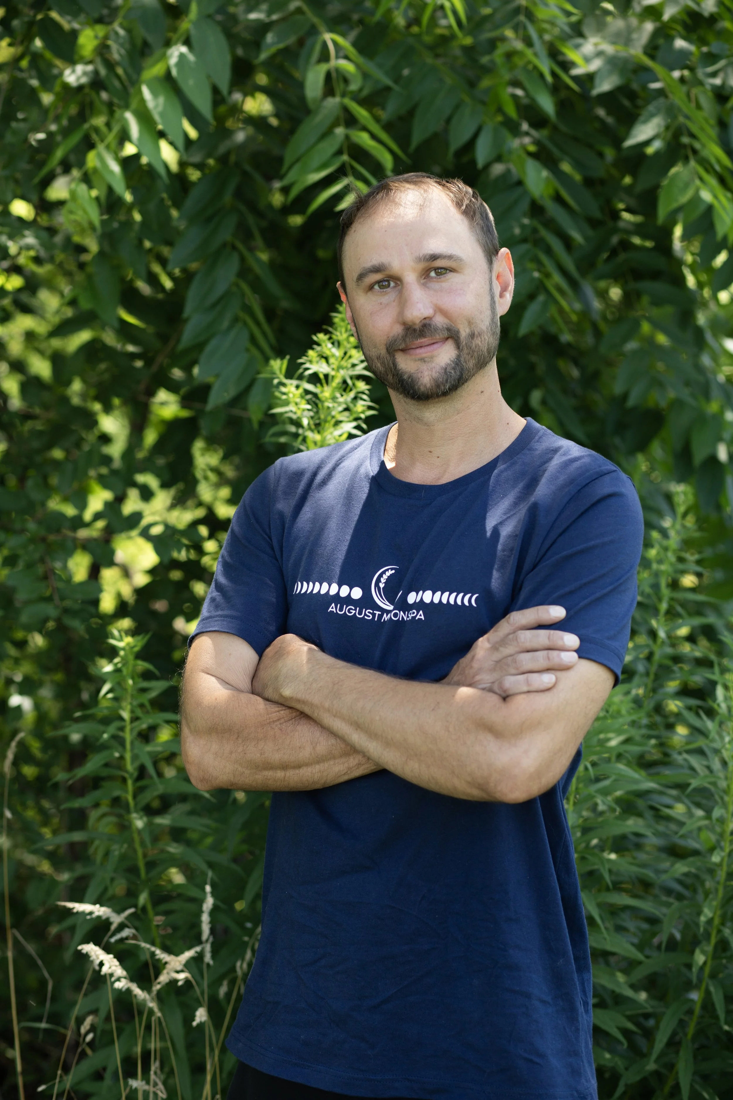 A smiling man with a beard and short hair standing outdoors in a natural setting with trees and grass.