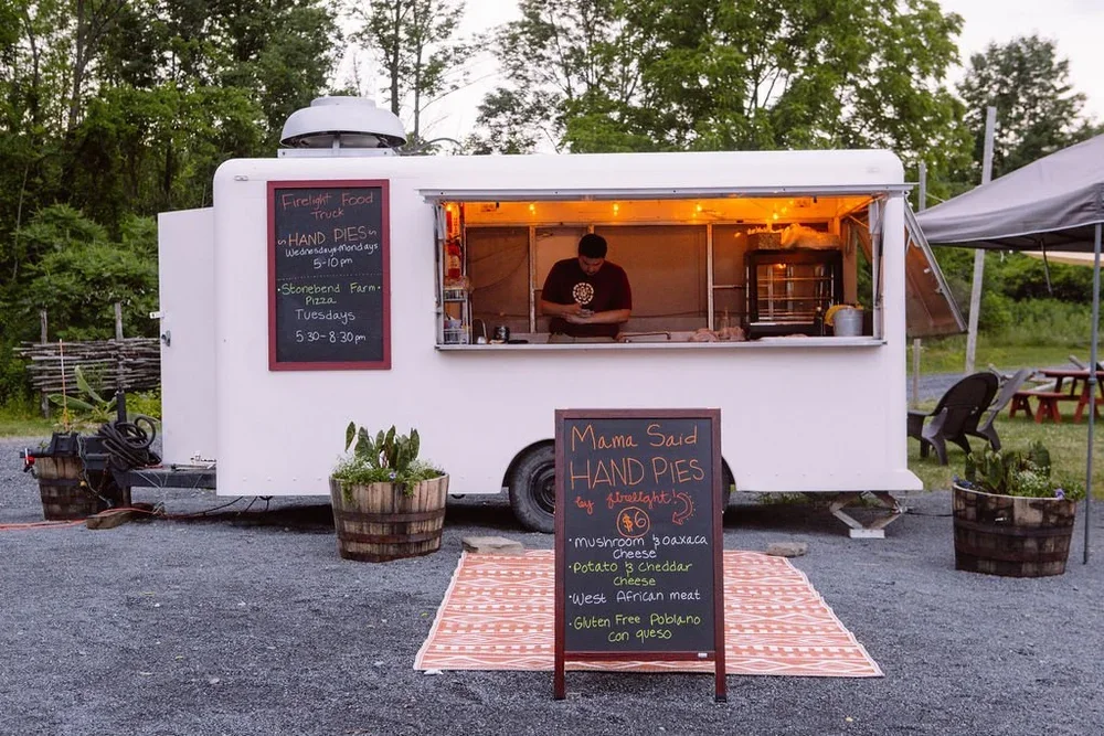 A white food truck is parked outdoors on a gravel area, selling handmade pies. The truck has a menu sign listing flavors including mushroom Oaxaca, potato cheddar, and West African meat. There is a man inside preparing food, and a blackboard sign outside displays additional menu details. The setup includes potted plants and an outdoor seating area with a canopy and picnic tables.