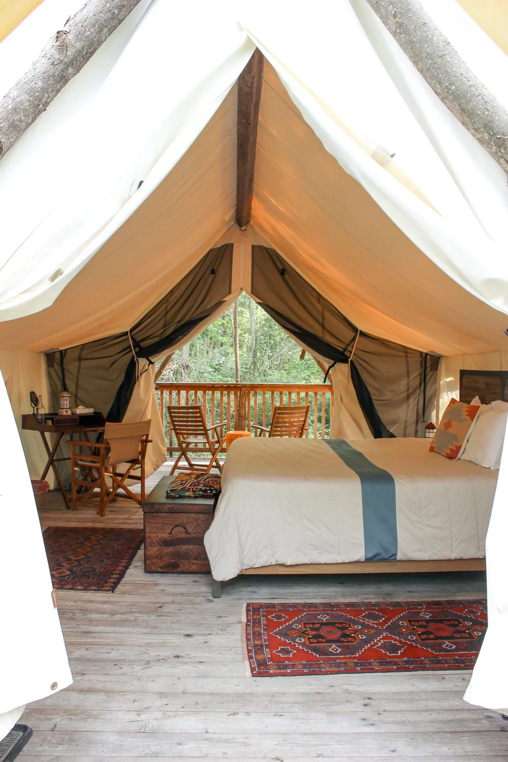 Interior of a cozy glamping tent with a bed, rug, small table, chairs, and a balcony overlooking trees.