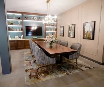 Dining room with wooden table, six upholstered chairs, a chandelier, a floral centerpiece, and built-in shelving with decorative items and a flat-screen TV.