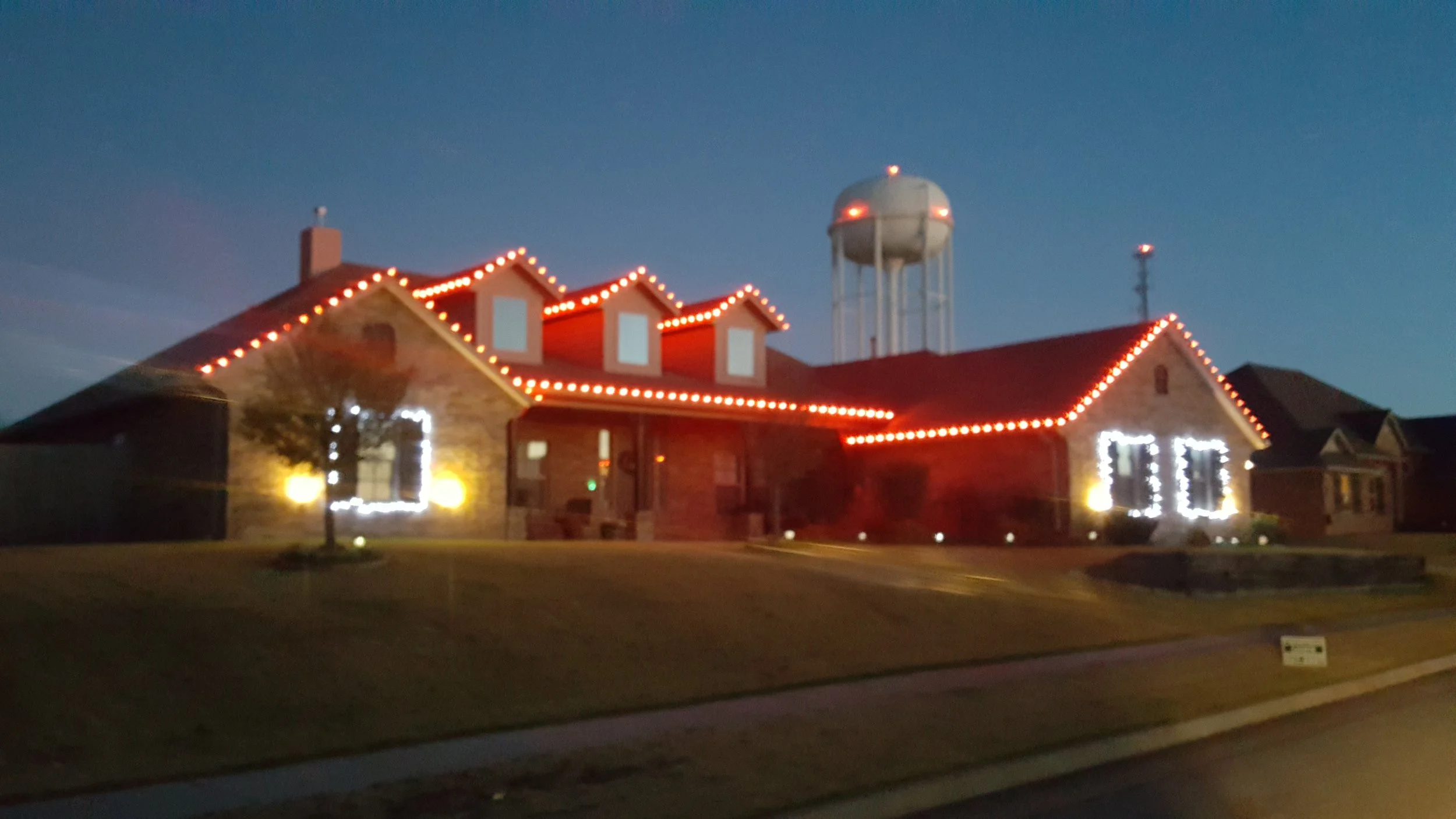 A house decorated with Christmas lights at dusk with a water tower in the background.
