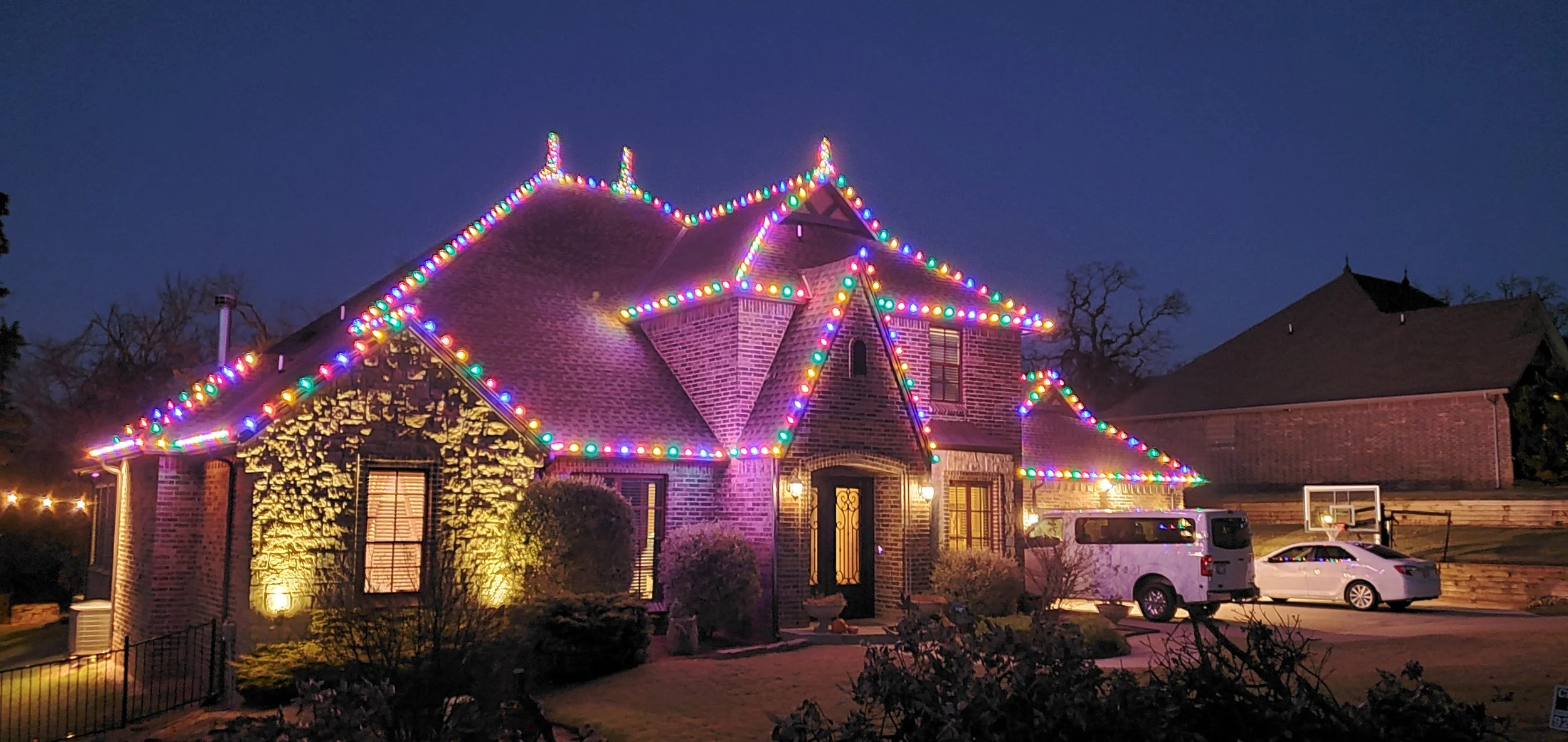 A brick house decorated with colorful Christmas lights at night, with a driveway and parked cars, a basketball hoop in the background, and leafless trees.