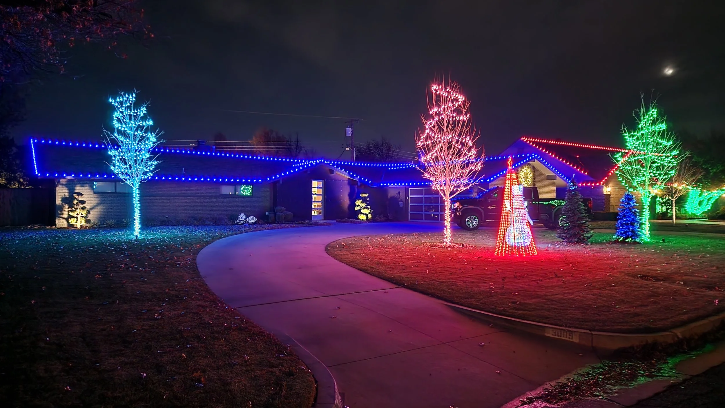 House decorated with colorful Christmas lights, including blue, red, and green lights on trees and house, with lit Christmas tree and snowman figure in the yard at night.