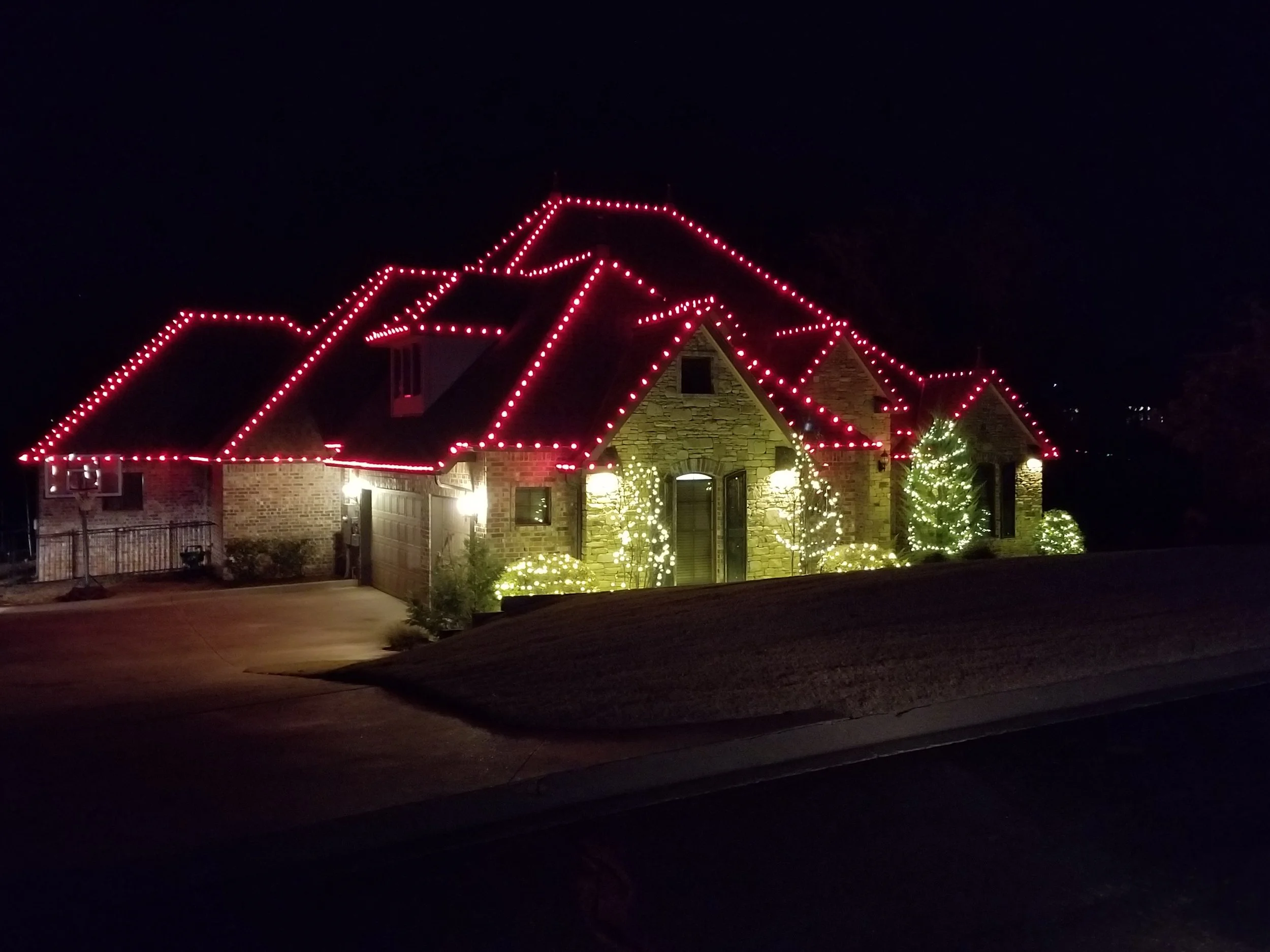 A house decorated with Christmas lights at night, featuring red lights outlining the roof and white lights on trees and bushes in front.