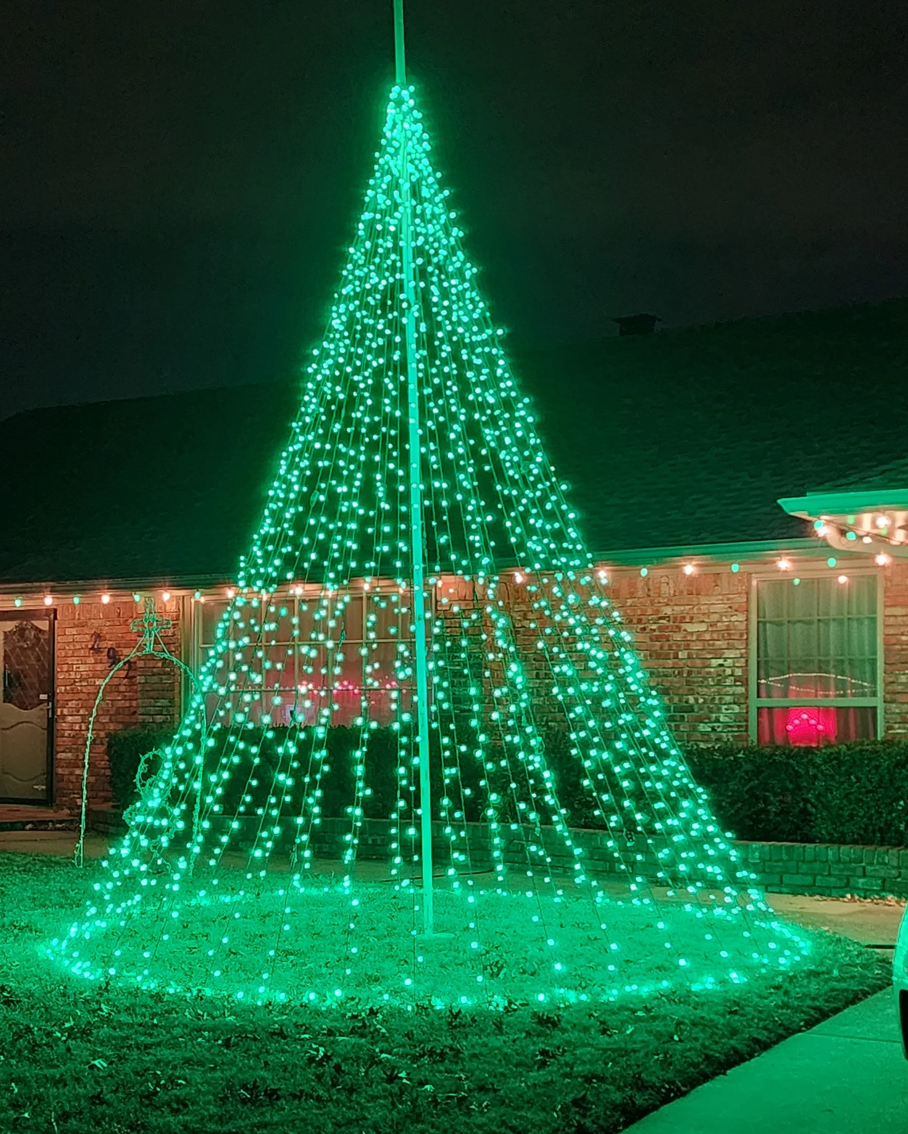 A house decorated with green Christmas lights arranged in the shape of a cone, resembling a Christmas tree, in the front yard at night.