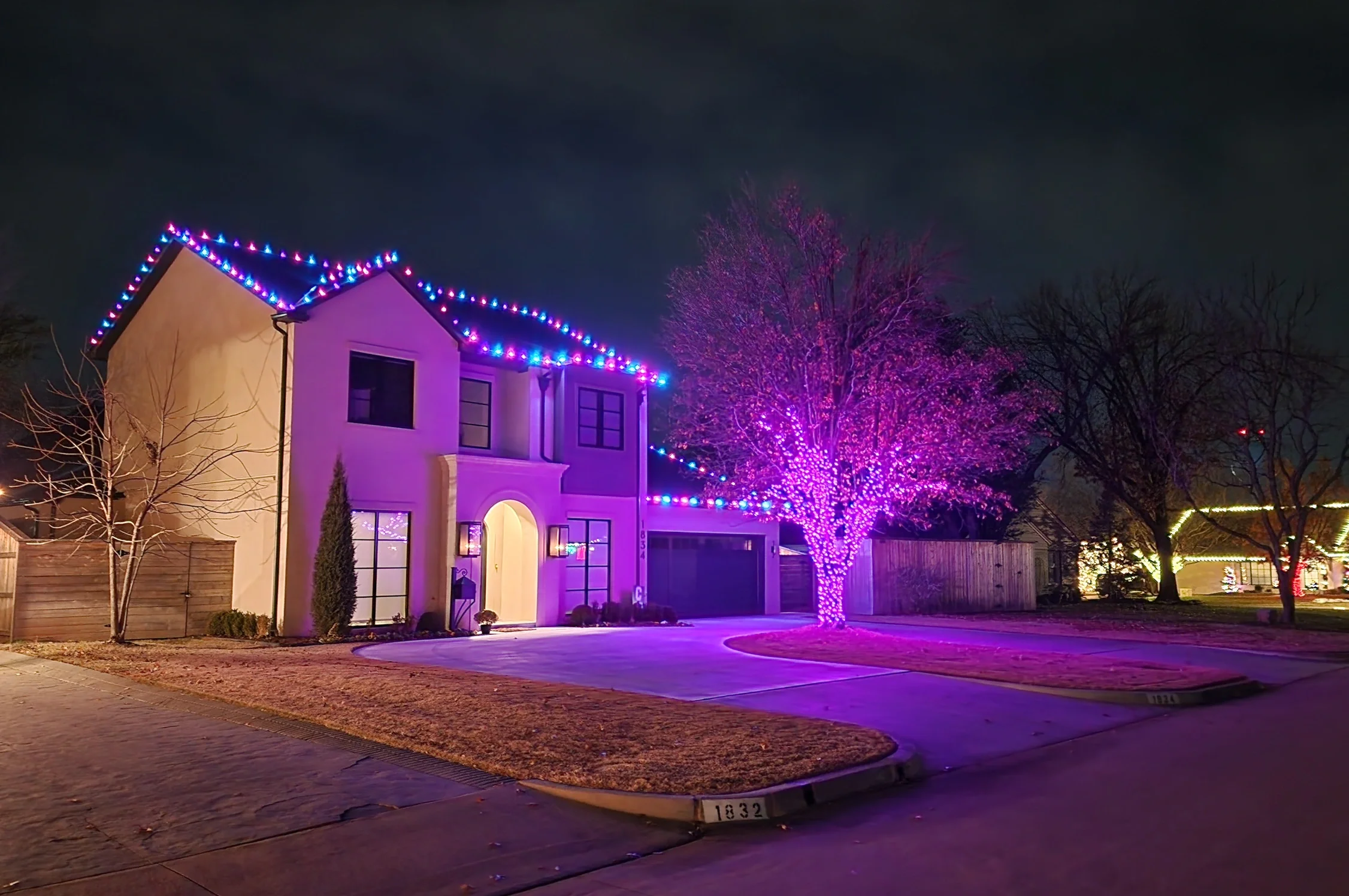 A house decorated with colorful Christmas lights at night, with a tree wrapped in purple lights and other trees with lights in the neighborhood.