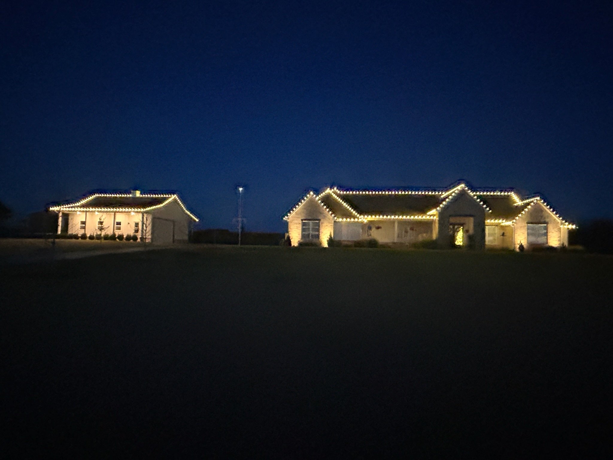 Large single-story house decorated with string lights along the roofline at night, with a smaller detached garage or outbuilding also illuminated.